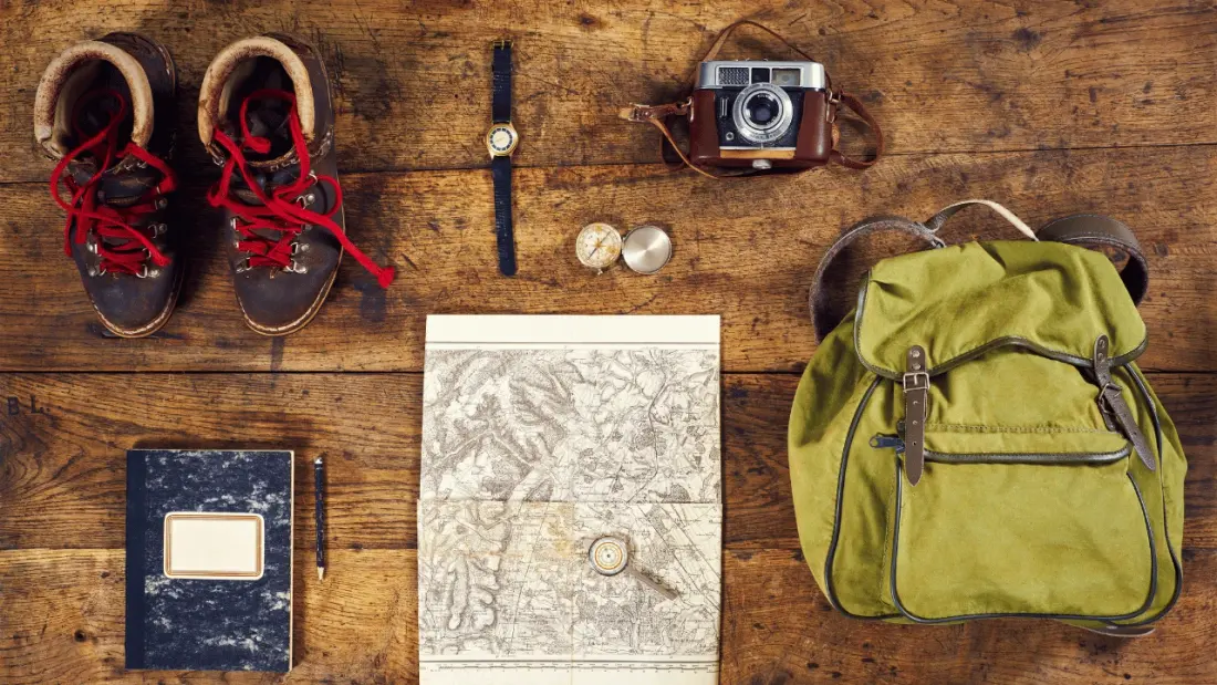 Trail gear laid out on a wooden table: hiking boots, a compass, a paper map, a notebook and a backpack